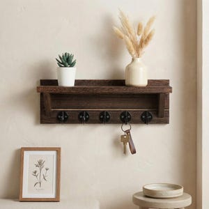 Op de afbeelding: Een donkerbruin houten wandplank met een klein plankje, vijf zwarte metalen haken en een kleine opbergruimte. De plank bevat een kleine potplant, een vaas met gedroogde bloemen en een sleutelbos.
