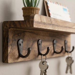 May include: A rustic wooden key holder with four metal hooks. The key holder has a small shelf and is stained a warm brown color. A small potted plant and an envelope are on the shelf. Keys hang from two of the hooks.