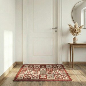 May include: A rectangular red and cream-colored patterned rug sits in front of a white door. The rug features a repeating geometric design with floral and snowflake-like motifs. The rug is on a wooden floor.