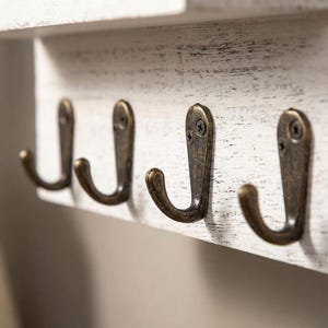 May include: A distressed white wooden wall-mounted shelf with four antique bronze metal hooks. The hooks are curved and attached with screws. The shelf has a rustic, farmhouse style.
