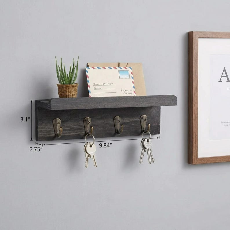 May include: A dark gray wooden key holder with four metal hooks and a small shelf. The key holder is 9.84 inches wide and 3.1 inches high. A small potted plant and an envelope are on the shelf. Keys hang from the hooks.