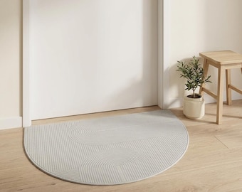 May include: A light gray, semi-circular doormat with a geometric design of curved lines. The mat is placed in front of a white door on a light wood floor. A small potted plant and a wooden stool are visible.