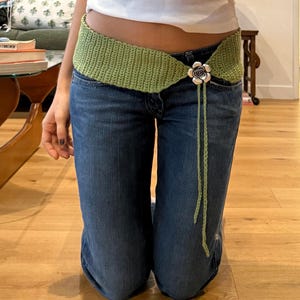 May include: A light green crochet belt with a floral buckle and long ties. The belt is worn over blue jeans. The floral buckle is silver and white. The belt is a unique fashion accessory.