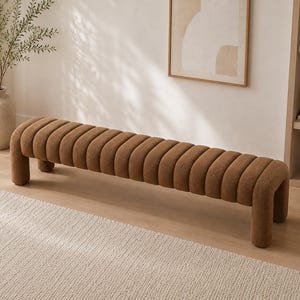 May include: A long, low-profile bench upholstered in a textured, brown fabric. The seat features a series of vertical, channel-tufted cushions that extend over the sides to form rounded, continuous legs at each end of the furniture piece.