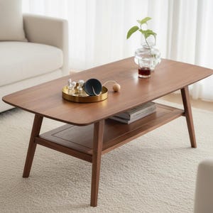 May include: A rectangular wooden coffee table with rounded corners and four angled legs. The table features a lower shelf holding two books. On the tabletop sits a round gold tray with small glass objects, a dark bowl, and a glass vase with a plant.