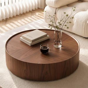 May include: A round, low-profile coffee table crafted from dark brown walnut wood with a visible chevron grain pattern. The table surface holds two stacked beige books, a clear glass vase with thin green branches, and a glass mug of black coffee.