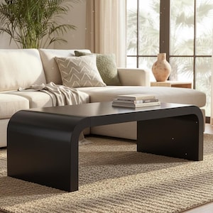 May include: A modern living room scene featuring a black coffee table with rounded edges. The table sits on a beige rug in front of a cream-colored sectional sofa with decorative pillows. A vase and books are on the table.