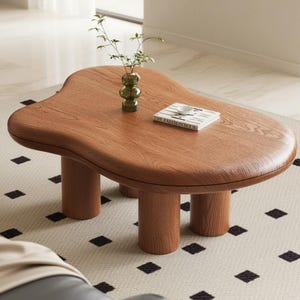 May include: A wooden coffee table with a unique, organic shape and four cylindrical legs. The table is a warm brown color and has a book and a small vase with greenery on top. The table sits on a patterned rug.