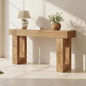 May include: A rectangular light brown wooden console table with thick, open-frame legs. On the table surface sits a beige lamp with a pleated shade and a woven basket containing green foliage, alongside a stack of two white books.