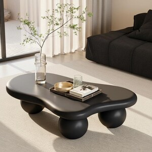 May include: A modern black coffee table with a unique, organic shape, supported by four rounded legs. A tray with a bowl, glass, and book sits on top. A clear vase with greenery is also present, adding a touch of nature to the room.