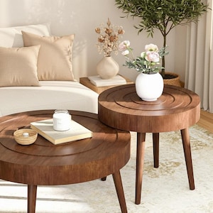 May include: Two round, dark brown wooden coffee tables with three legs each. The tables are nested together. A white vase with flowers sits on the smaller table. A book, candle, and small bowl are on the larger table.