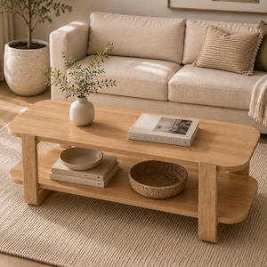 May include: A light-toned wooden coffee table with a rectangular top and a lower shelf. The top surface holds a white vase with green foliage and a book titled Live Beautifully. The lower shelf holds two stacked books and a woven bowl.