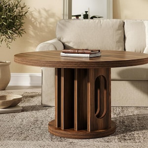 May include: A round, dark brown wooden coffee table with a unique base design. The base features vertical slats and an oval cutout. A book rests on the table's surface. The table is in a living room setting with a sofa and a potted plant.