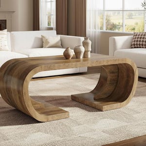 May include: A modern, curved wooden coffee table with a warm brown finish. The table has a unique, wave-like design and sits on a patterned rug. Decorative vases are placed on the table, and white sofas are in the background.