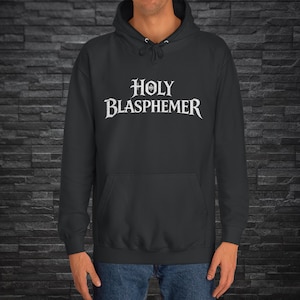 Puede incluir: Sudadera con capucha gris oscuro con el texto blanco "HOLY BLASPHEMER" en letras góticas. La sudadera tiene una capucha con cordón y un bolsillo delantero. La persona lleva vaqueros azules.