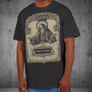 Puede incluir: Camiseta gris oscuro con un diseño vintage. Presenta una figura esquelética en una pose religiosa, con las palabras "GOD" y "I DON'T EXIST" incorporadas en la obra de arte. El diseño incluye nubes y ángeles.