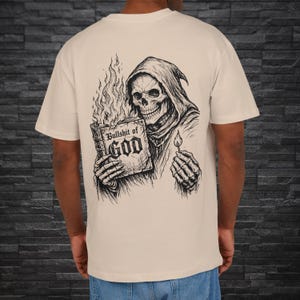 Puede incluir: Camiseta color crema con una ilustración detallada en blanco y negro de una figura esquelética sosteniendo un libro en llamas con el texto "Bullshit of GOD". El diseño incluye una calavera con capucha y un fósforo encendido.