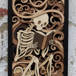May include: A wooden wall art piece featuring a skeleton sitting and reading a book. The skeleton is surrounded by intricate, carved swirls and patterns in shades of brown.