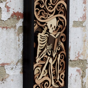 May include: A wooden wall art piece featuring a skeleton reading a book. The skeleton is surrounded by intricate, layered swirls and designs, creating a 3D effect. The piece is framed in black.