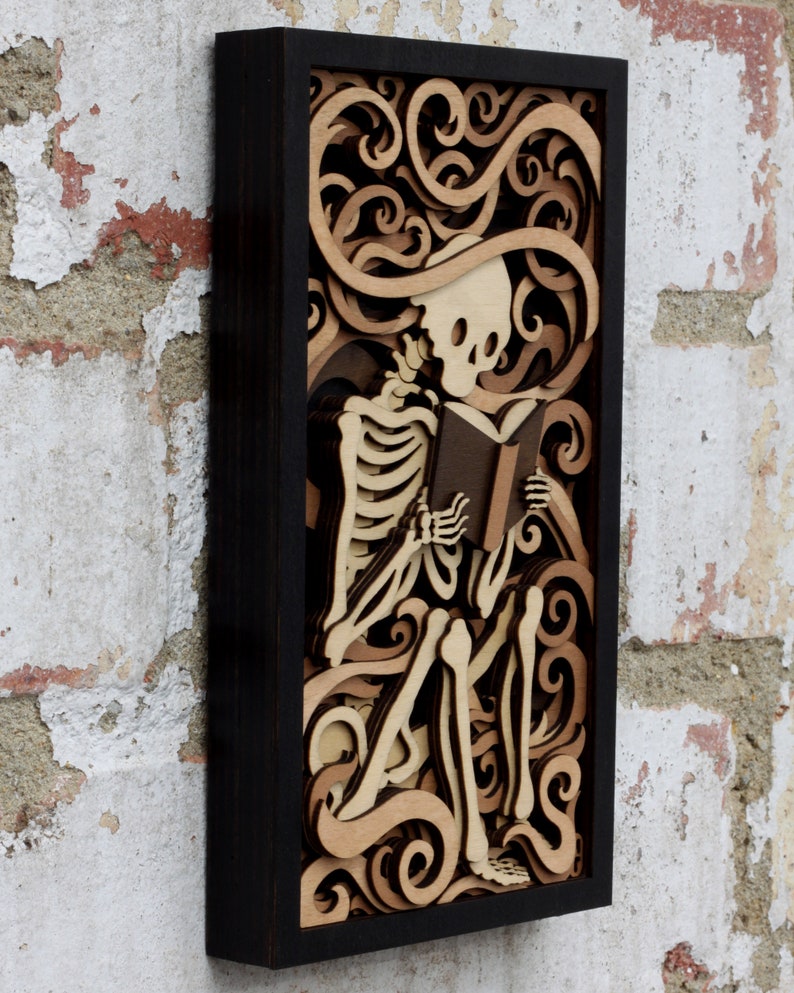 May include: A wooden wall art piece featuring a skeleton reading a book. The skeleton is surrounded by intricate, layered swirls and designs, creating a 3D effect. The piece is framed in black.