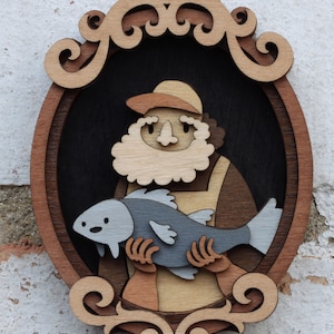 Puede incluir: Una obra de arte de madera en capas que representa a un pescador sosteniendo un pez. La obra presenta a un pescador de piel clara con barba y sombrero, sosteniendo un pez gris. La pieza est&aacute; enmarcada en una forma ovalada decorativa con un borde marr&oacute;n claro.