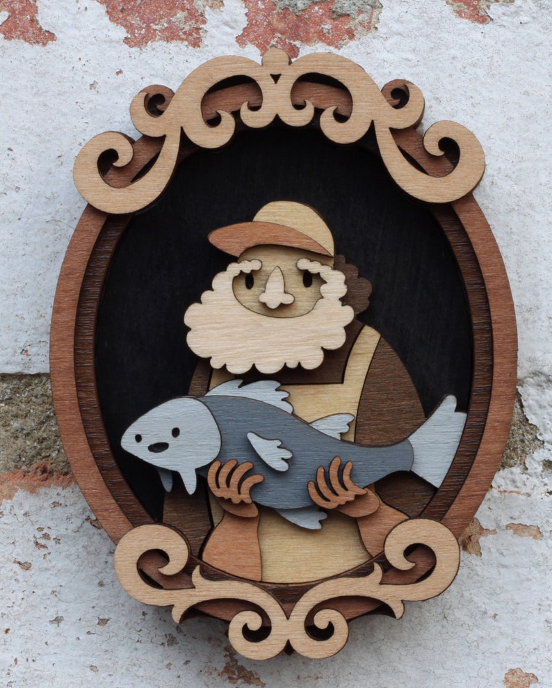 Puede incluir: Una obra de arte de madera en capas que representa a un pescador sosteniendo un pez. La obra presenta a un pescador de piel clara con barba y sombrero, sosteniendo un pez gris. La pieza est&aacute; enmarcada en una forma ovalada decorativa con un borde marr&oacute;n claro.