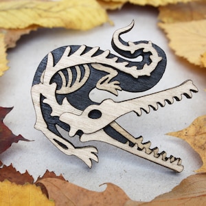 May include: A wooden alligator skeleton pin with a black background. The alligator is in a running pose with its mouth open and teeth visible. The pin is made of wood and has a natural wood finish.