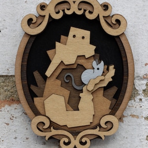 May include: A wooden wall hanging with a decorative frame. The frame is oval shaped and has a brown wood finish. The center of the frame features a cutout design of a brown and gray mouse climbing a brown structure.