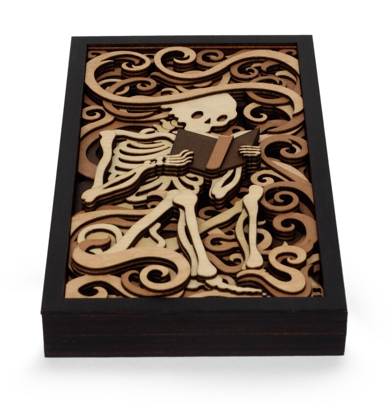 May include: A wooden wall art piece featuring a skeleton reading a book. The skeleton is surrounded by intricate swirls and patterns, creating a whimsical and spooky design.