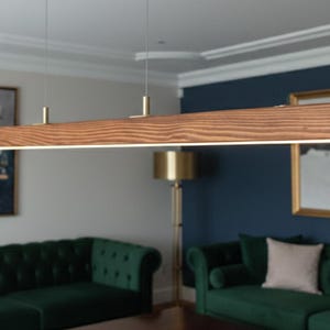 Può includere: Una moderna lampada a sospensione rettangolare in legno con finitura naturale e calda. La lampada ha una striscia LED integrata ed è sospesa al soffitto con sottili fili. È posizionata sopra un tavolino in legno scuro e divani in velluto verde.