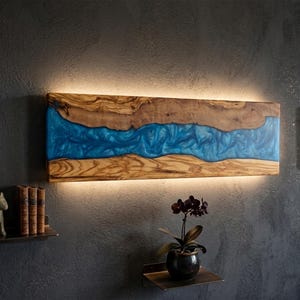 Op de afbeelding: Een rechthoekige wandlamp bestaande uit een paneel van natuurlijk hout met een centrale, golvende blauwe harsinleg. Het object wordt van achteren verlicht, werpt een zachte gloed op de muur en creëert een rivierachtig visueel effect.
