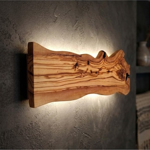 May include: A rectangular wall-mounted light fixture crafted from a single piece of natural olive wood with a live edge. The wood features prominent brown grain patterns and knots, with integrated LED lighting casting a soft glow on the wall.