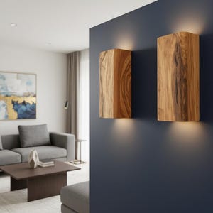 May include: Two rectangular wall sconces crafted from natural wood with a prominent grain pattern. Each fixture is mounted on a dark blue wall and emits a warm, soft glow from both the top and bottom edges, creating an ambient lighting effect.