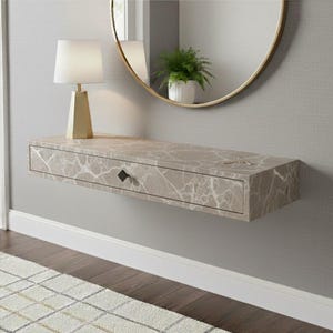 Op de afbeelding: Een zwevende console met marmerlook, met een enkele lade en een zwarte, ruitvormige handgreep. Op de tafel staat een goudkleurige lamp. Een ronde spiegel en een kleine plant zijn zichtbaar op de achtergrond.