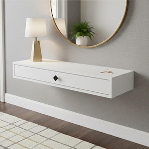 Puede incluir: Un cajón blanco flotante montado en la pared con un tirador negro en forma de diamante. Una lámpara dorada y una pequeña planta en maceta encima. Un espejo con marco dorado cuelga de la pared. Llaves sobre el cajón.