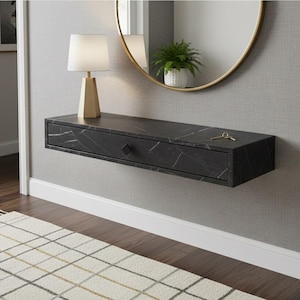 Pode incluir: Uma consola flutuante preta com efeito mármore, uma gaveta e uma chave dourada. Uma lâmpada dourada com abajur branco e um espelho redondo dourado estão montados acima da mesa. Uma pequena planta em vaso é visível.