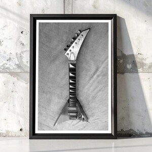 Może przedstawiać: Czarno-biała oprawiona grafika przedstawiająca gitarę elektryczną. Gitara ma korpus w kształcie litery V i gł&oacute;wkę z kołkami do strojenia. Widoczna jest podstrunnica i struny. Grafika jest na tle teksturowanego szarego tła.