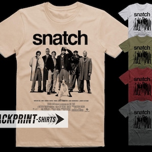 Op de afbeelding: Een beige t-shirt met korte mouwen voorzien van de filmtitel Snatch in vetgedrukte zwarte letters boven een monochrome castfoto en een hond. Aan de rechterkant worden kleinere t-shirts in wit, olijfgroen, bordeauxrood en donkergrijs weergegeven.