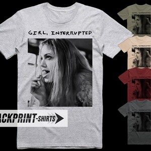 Op de afbeelding: Een lichtgrijs t-shirt met korte mouwen voorzien van een zwart-wit fotoprint van een persoon met lang haar en de tekst "GIRL, INTERRUPTED" boven de afbeelding. Het ontwerp is ook getoond in olijf, beige, bordeaux en donkergrijs.