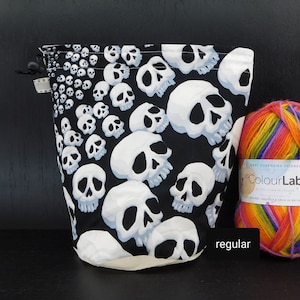 Könnte beinhalten: Eine weiße Kordeltasche mit schwarzem Hintergrund und einem Totenkopf-Muster. Die Tasche ist aus Stoff und hat eine weiße Kordel. Die Tasche liegt auf einer schwarzen Oberfläche neben einem Knäuel Regenbogenwolle.