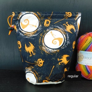 Könnte beinhalten: Eine schwarze Kordeltasche mit einem weißen und orangefarbenen Druck, der einen Charakter aus Nightmare Before Christmas zeigt. Der Druck enthält auch Kürbisse und Fledermäuse. Die Tasche liegt auf einer schwarzen Oberfläche neben einem Knäuel Regenbogenwolle.
