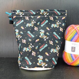 Puede incluir: Bolsa de cordón negra con forro blanco. La bolsa está impresa con un patrón repetido de un personaje de dibujos animados en un traje espacial con las palabras "Greetings Earthlings" en una fuente retro.