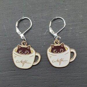 Op de afbeelding: Een paar gouden oorbellen met een witte koffiemok met een zwarte kat die uit de bovenkant kijkt. De tekst "Caffe" staat op de zijkant van de mok.