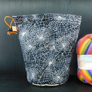 Puede incluir: Una bolsa de cordón negro con un patrón de telaraña blanca y abejas doradas. La bolsa está hecha de tela y tiene un forro blanco. La bolsa está sentada sobre una superficie negra junto a una bola de hilo de arcoíris.