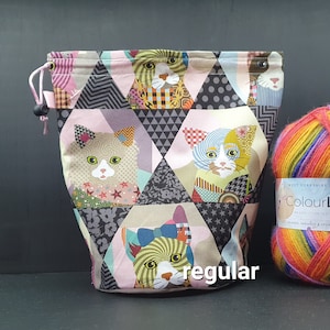 May include: A pink and grey drawstring bag with a patchwork design featuring cats. The bag is made of fabric and has a drawstring closure. The bag is sitting on a black surface next to a ball of colourful yarn.