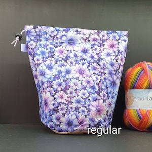 Puede incluir: Una bolsa de proyecto de cordón morado con un estampado floral de flores blancas y moradas. La bolsa está sentada junto a una bola de hilo arcoíris.