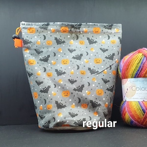 May include: A grey drawstring bag with a Halloween print of orange pumpkins, black bats, white stars, and candy corn. The bag is sitting on a table next to a ball of rainbow yarn.