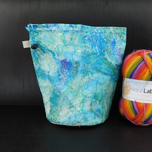 May include: A blue and green fabric drawstring bag with a white marble pattern. The bag is sitting on a black surface next to a ball of rainbow colored yarn.