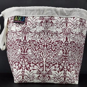 May include: A burgundy drawstring bag with a white floral pattern featuring birds and rabbits. The bag has a white cotton lining and a label that reads "AXJ amellajoy".