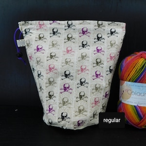 May include: A white drawstring bag with a skull and crossbones pattern. The bag is made of fabric and has a purple drawstring. The bag is sitting on a black surface next to a ball of yarn.